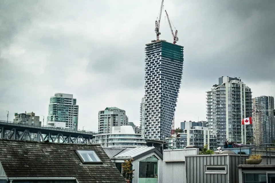View of Vancouver House skycraper under construction with two cranes at top and other downtown Vancouver buildings surrounding it against a grey sky